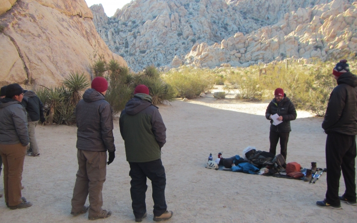 A group of people stand in the shade of large rock wall and appear to be listening to an instructor give a lesson on backpacking. There is another large rock wall in the background. 